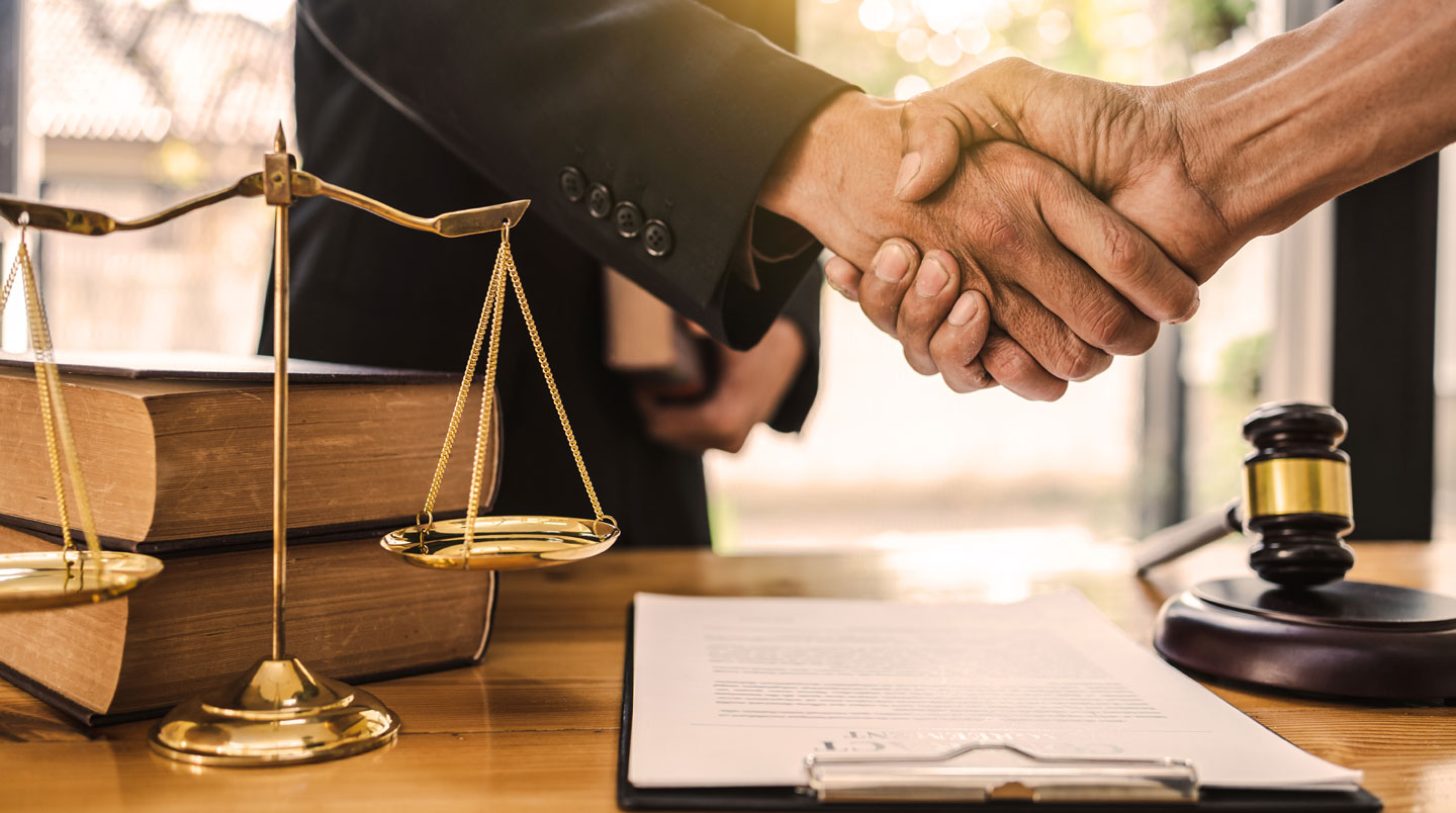 Mains gantées tenant une balance de justice miniature sous une lumière focalisée, avec une bibliothèque juridique en arrière-plan flou.